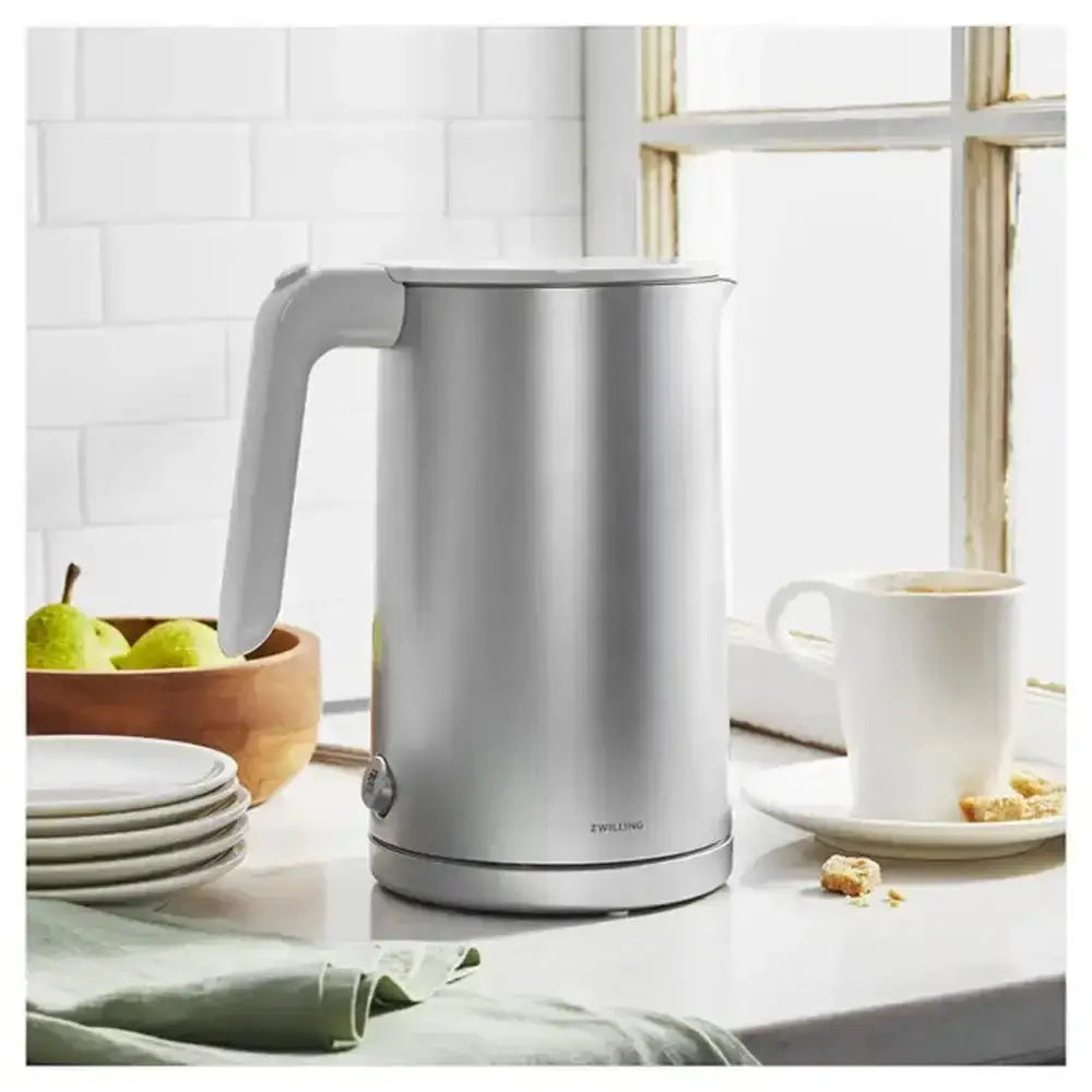 Silver electric kettle on a kitchen counter with a cup and fruit bowl in the background.