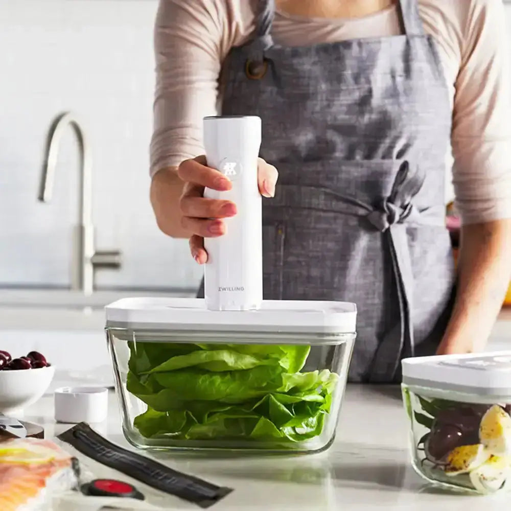 Person holding a white kitchen tool over a container of greens on a kitchen counter.