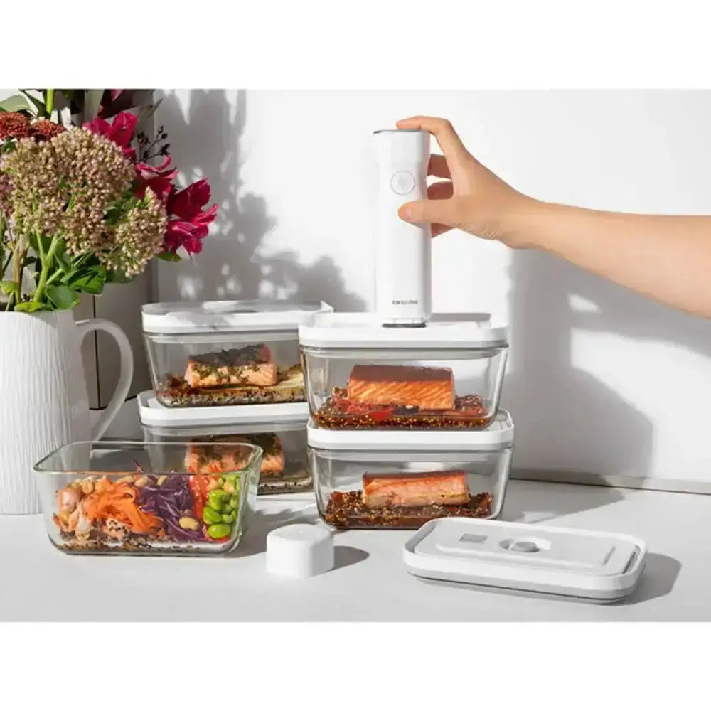 Hand using a food preservation device on stacked containers with food, next to flowers and a mug.