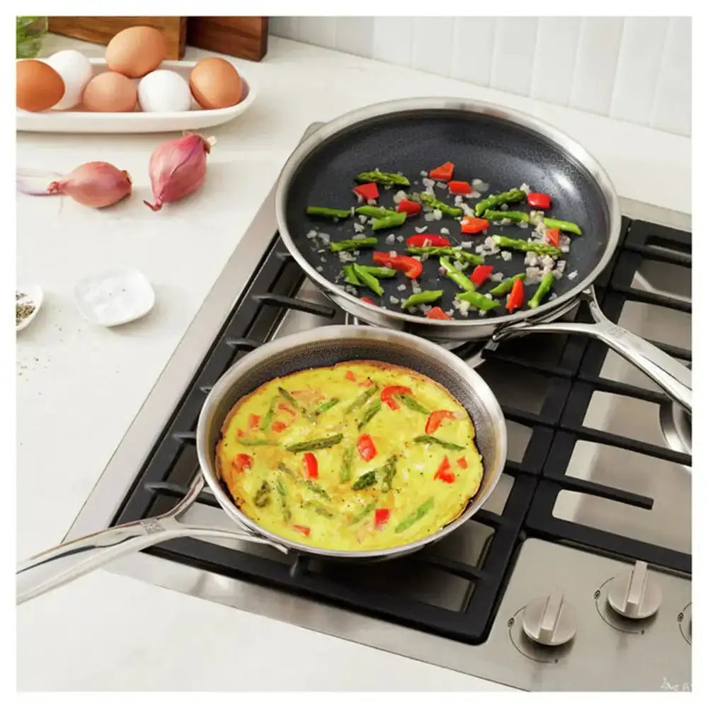 Two frying pans on a stove with vegetables and a frittata, surrounded by eggs and shallots on a kitchen counter.
