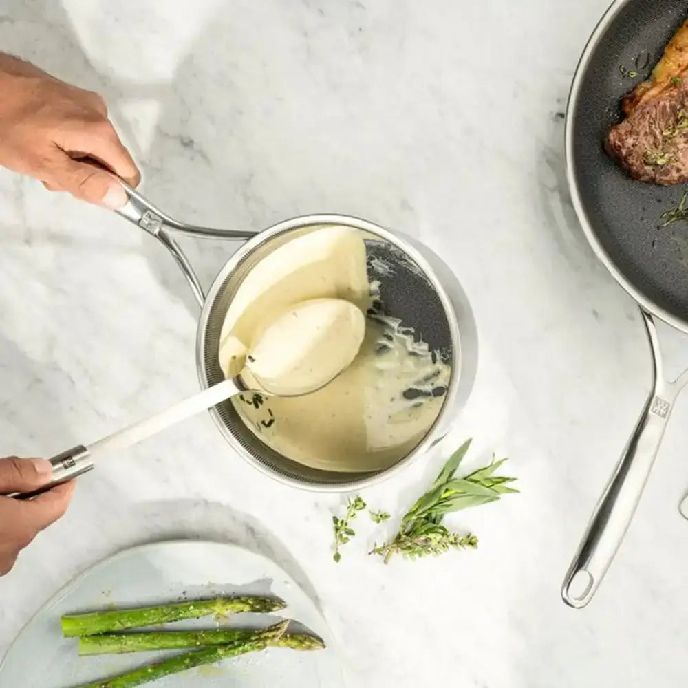 Person ladling creamy sauce from a small pot onto a plate with asparagus on a marble surface.
