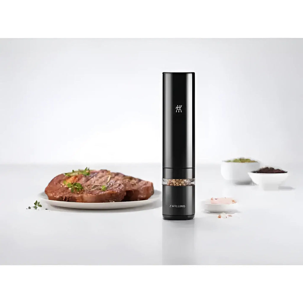 Black pepper grinder on a white surface with cooked meat and small bowls of spices in the background.