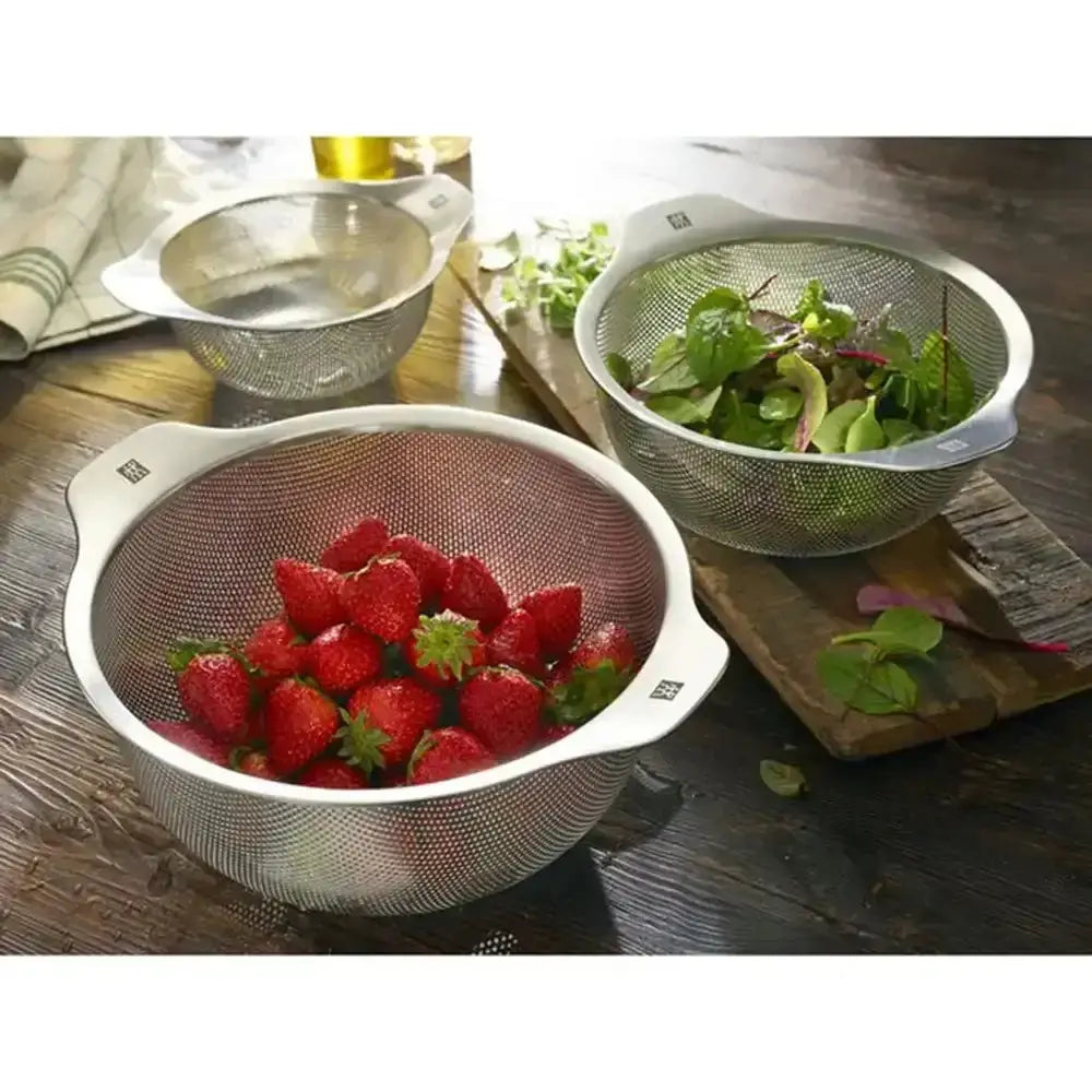 Two colanders on a wooden surface, one with strawberries and the other with salad greens.