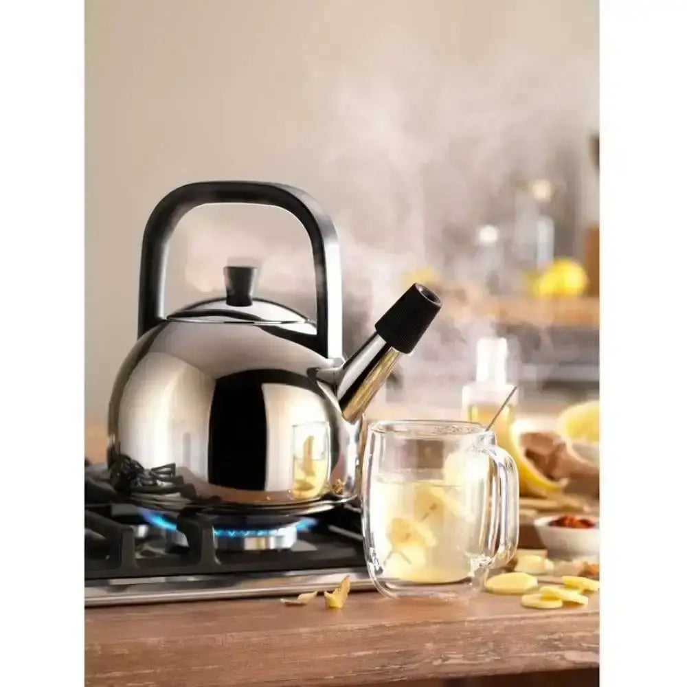 Stainless steel kettle on a stove with steam rising, next to a glass mug with lemon slices.