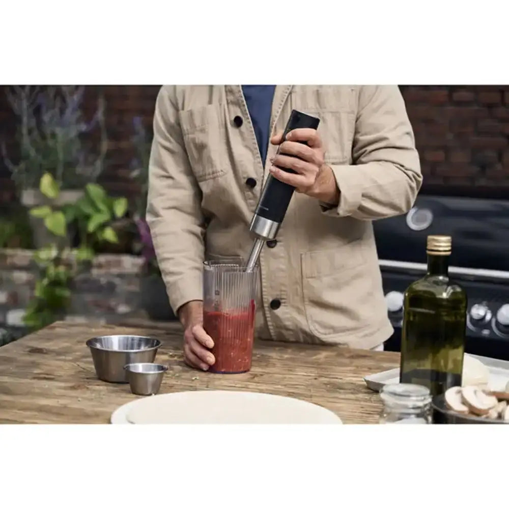 Person using a handheld blender with a container on a wooden table outdoors.