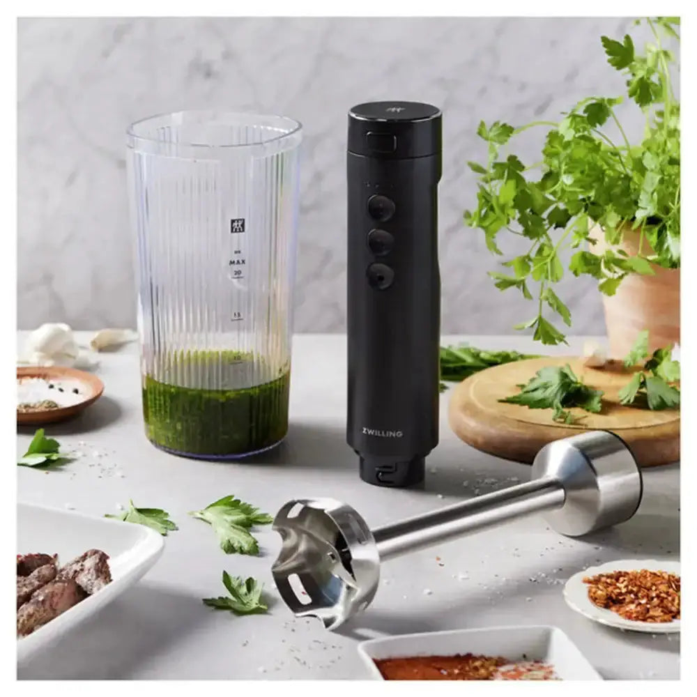 Black immersion blender with clear container on a kitchen counter with ingredients and a plant.