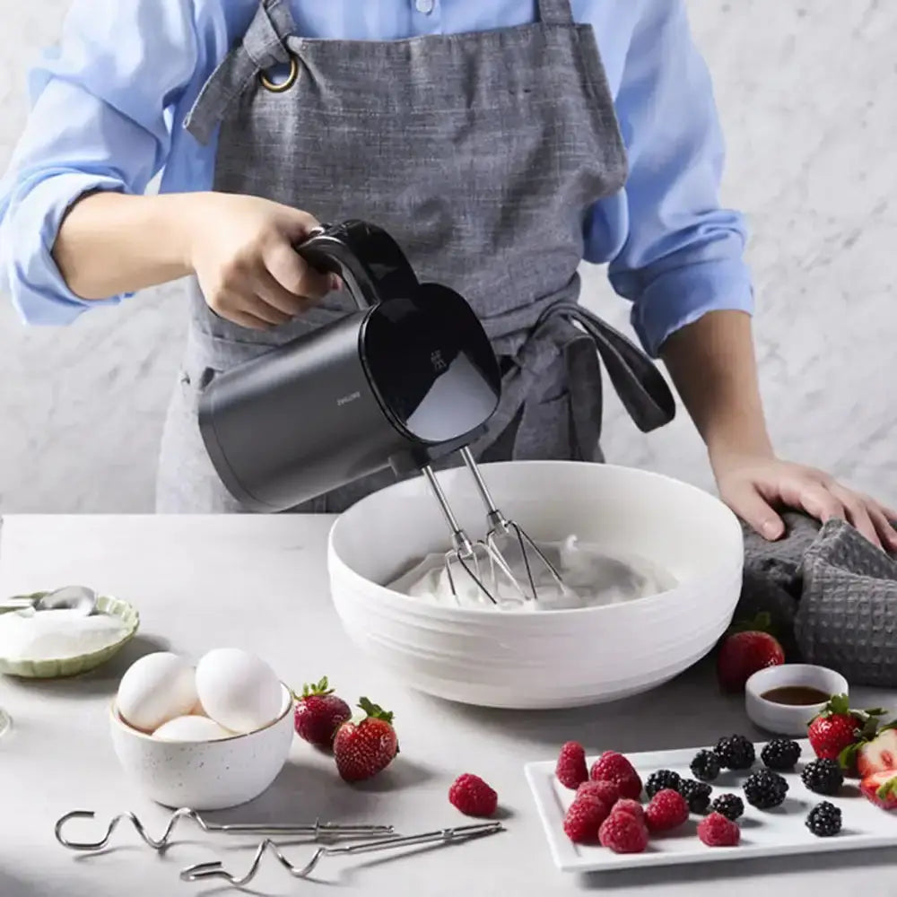 Person using a hand mixer in a kitchen with ingredients and utensils around.